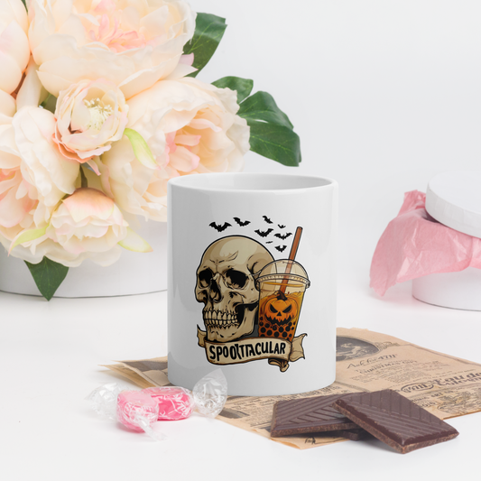 White mug with skull design and 'Spooktacular' text, surrounded by flowers and treats on a white surface.